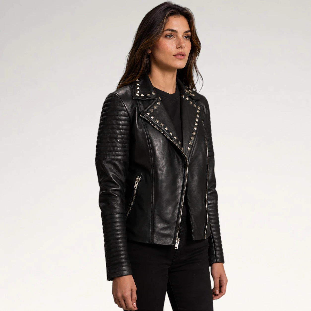A woman modeling a black faux-leather moto jacket with silver studs and quilted sleeve panels, paired with a black top and black pants, against a light gray studio background.
