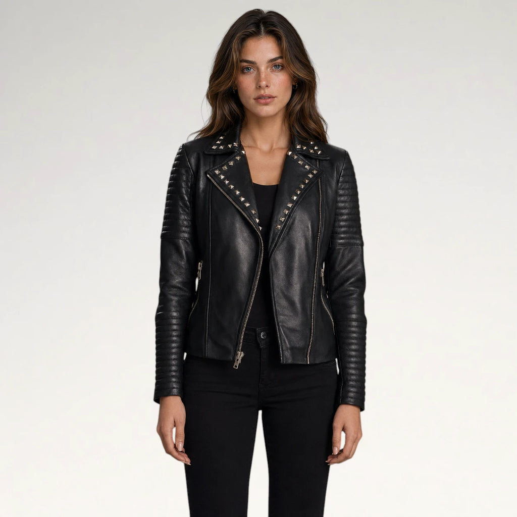 A woman modeling a black faux-leather moto jacket with silver studs and zippers, paired with a black top and black pants, against a light gray studio background.