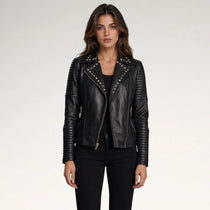 A woman modeling a black faux-leather moto jacket with silver studs and zippers, paired with a black top and black pants, against a light gray studio background.