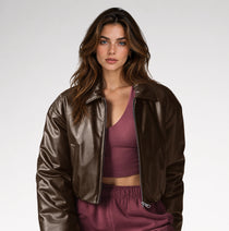 A woman modeling a cropped, shiny dark brown faux-leather jacket over a mauve-burgundy crop top and matching sweatpants, shot against a clean light gray studio background.