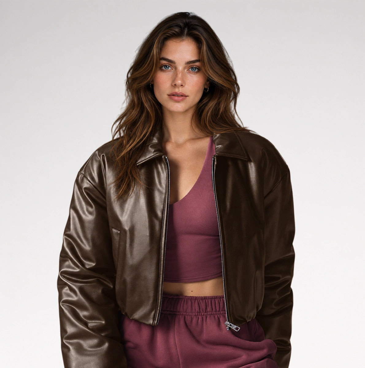 A woman modeling a cropped, shiny dark brown faux-leather jacket over a mauve-burgundy crop top and matching sweatpants, shot against a clean light gray studio background.