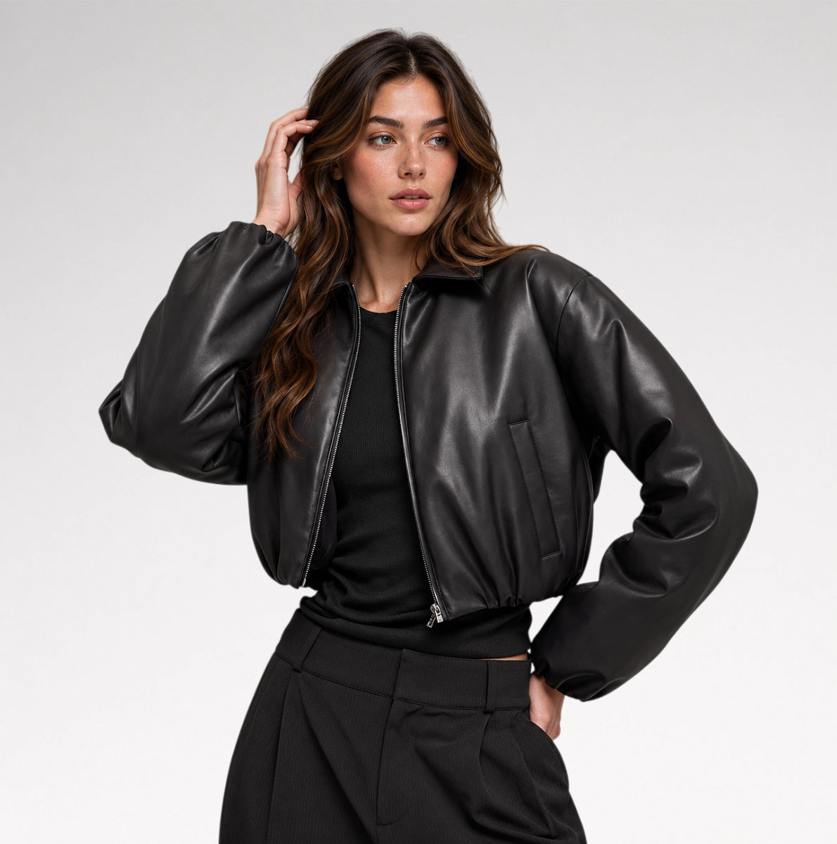 A woman modeling a black faux-leather cropped bomber jacket over a black top and black trousers, photographed against a light gray studio background. Main image