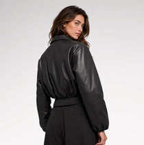 A woman wearing a black faux-leather jacket with balloon sleeves and a matching black belt, shown from a three-quarter back view against a light gray studio background.
