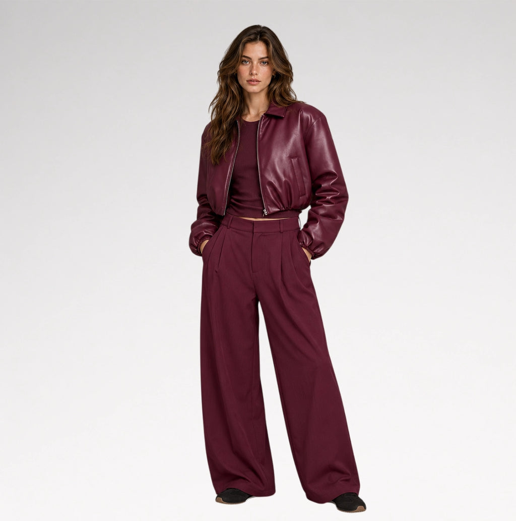 A woman wearing a glossy, deep burgundy faux-leather cropped jacket and matching trousers, photographed from the side against a clean white studio background.