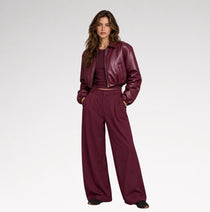 A woman wearing a glossy, deep burgundy faux-leather cropped jacket and matching trousers, photographed from the side against a clean white studio background.