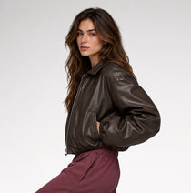 A woman wearing a dark brown faux-leather bomber jacket and burgundy sweatpants, photographed against a clean white studio background.