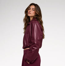 A woman wearing a glossy, deep burgundy faux-leather cropped jacket and matching trousers, photographed from the side against a clean white studio background.