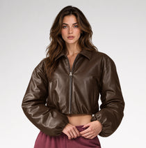 A woman modeling a cropped brown faux-leather jacket with a half-zip front, standing against a light gray studio background.