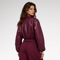 A woman wearing a glossy, deep burgundy faux-leather cropped jacket and matching trousers, photographed from the side against a clean white studio background.