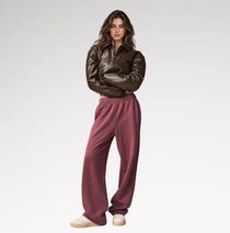 A model stands against a light gray studio background wearing a dark brown faux-leather cropped jacket and dusty rose wide-leg sweatpants.