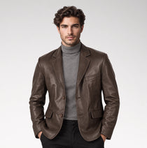 A man wearing a brown leather blazer over a gray turtleneck sweater and black trousers, standing against a light gray studio background.