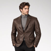 A man wearing a dark brown leather blazer over a gray turtleneck sweater and black pants, standing against a light gray studio background.