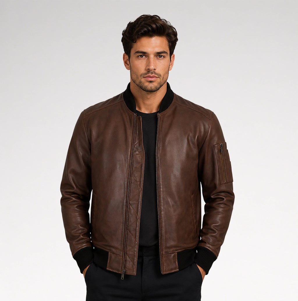 A man wearing a brown leather bomber jacket with black ribbed trim over a black shirt, standing against a light gray studio background.