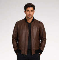 A man wearing a brown leather bomber jacket with black ribbed trim over a black shirt, standing against a light gray studio background.