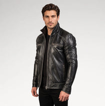 A man wearing a black leather motorcycle jacket with silver zippers, styled over a black shirt and black pants, photographed against a light gray studio background.