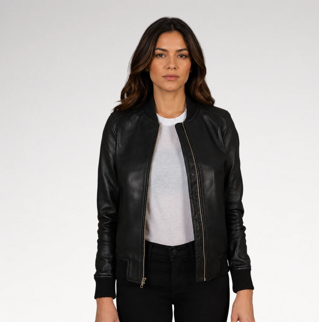 A woman modeling a black leather bomber jacket with gold-tone zippers, worn open over a light gray t-shirt and black pants, against a plain light gray studio background.