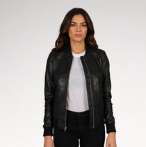 A woman modeling a black leather bomber jacket with gold-tone zippers, worn open over a light gray t-shirt and black pants, against a plain light gray studio background.