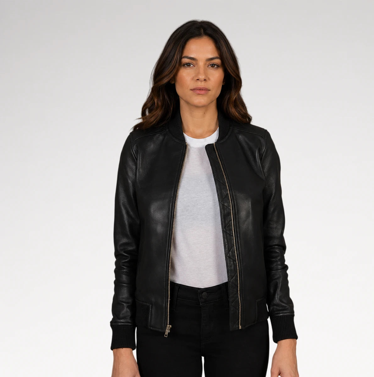 A woman modeling a black leather bomber jacket with gold-tone zippers, worn open over a light gray t-shirt and black pants, against a plain light gray studio background.