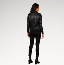 A woman viewed from the back wearing a black faux-leather jacket, black skinny pants, and black ankle boots against a light gray studio background.