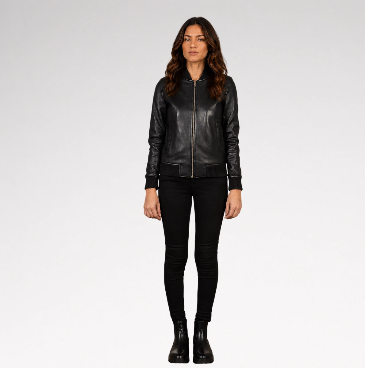 A woman modeling a black leather bomber jacket with a front zipper, paired with black skinny pants and black ankle boots, against a light gray studio background.