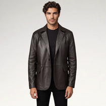 A man wearing a dark brown leather blazer over a black t-shirt and black pants, standing against a light gray studio background.