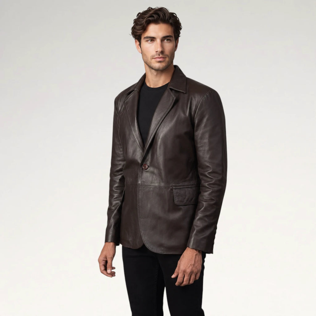 A man wearing a dark brown leather blazer over a black t-shirt and black pants, standing against a light gray studio background.
