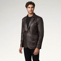A man wearing a dark brown leather blazer over a black t-shirt and black pants, standing against a light gray studio background.