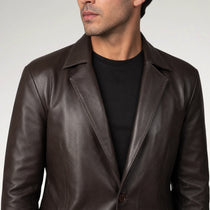 A man wearing a dark brown leather blazer over a black crewneck t-shirt, shown from the chest up against a light gray studio background.