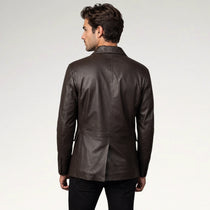 A man viewed from the back wearing a dark brown leather blazer over black trousers, set against a plain light gray studio background.