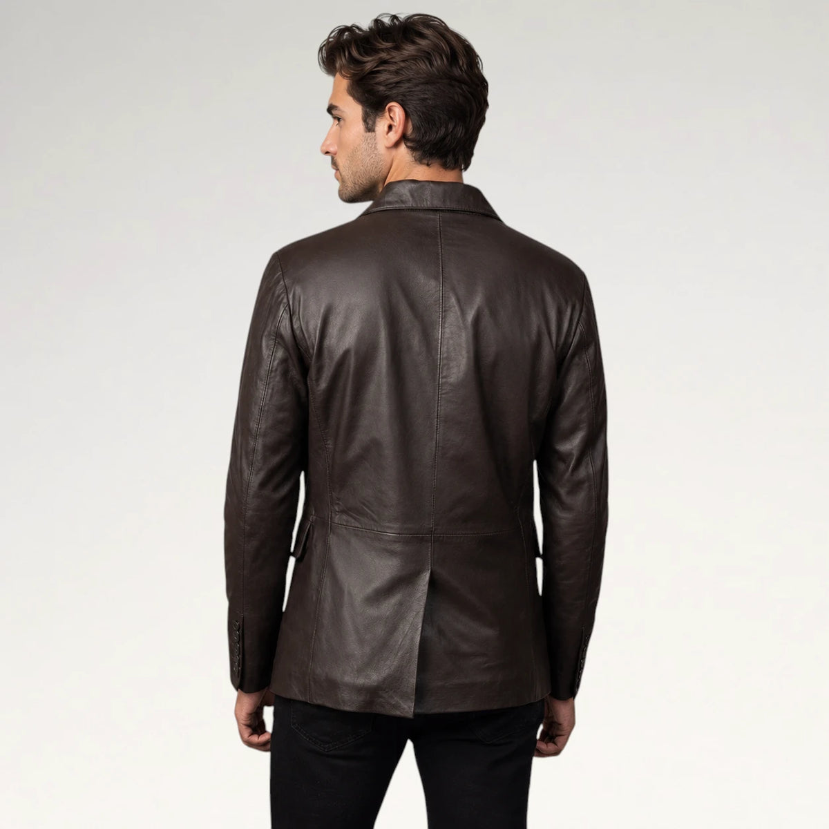 A man viewed from the back wearing a dark brown leather blazer over black trousers, set against a plain light gray studio background.