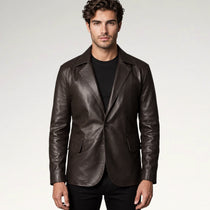 A man wearing a dark brown leather blazer over a black t-shirt and black pants, standing against a light gray studio background.