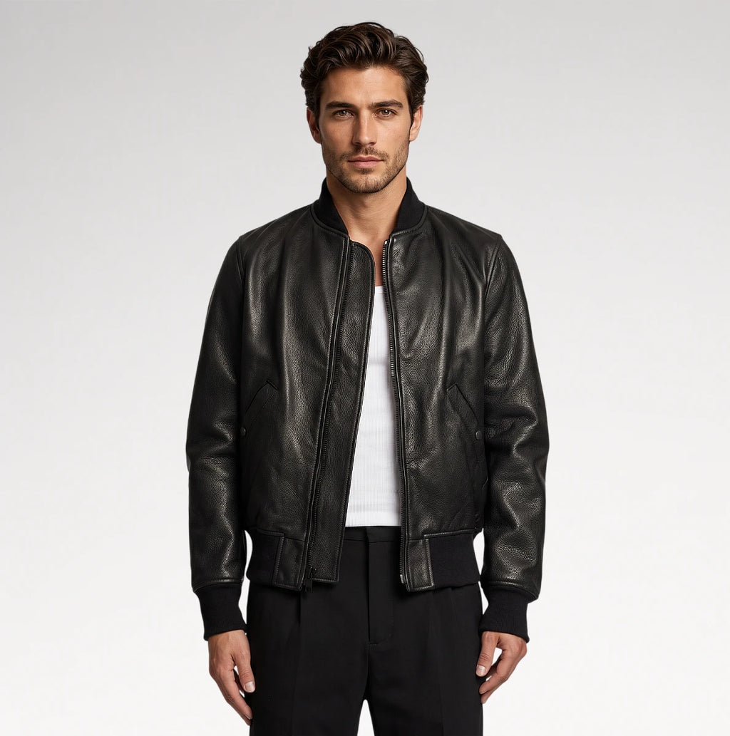 A man wearing a black leather bomber jacket over a white t-shirt and black pants, standing against a light gray studio background.