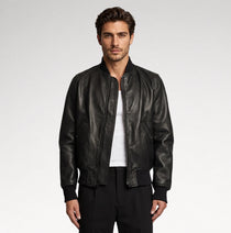 A man wearing a black leather bomber jacket over a white t-shirt and black pants, standing against a light gray studio background.