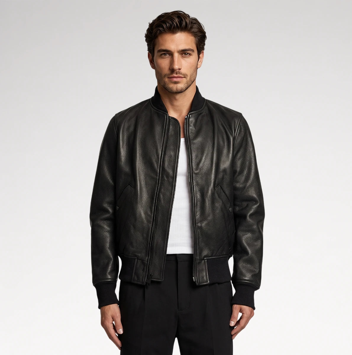 A man wearing a black leather bomber jacket over a white t-shirt and black pants, standing against a light gray studio background.