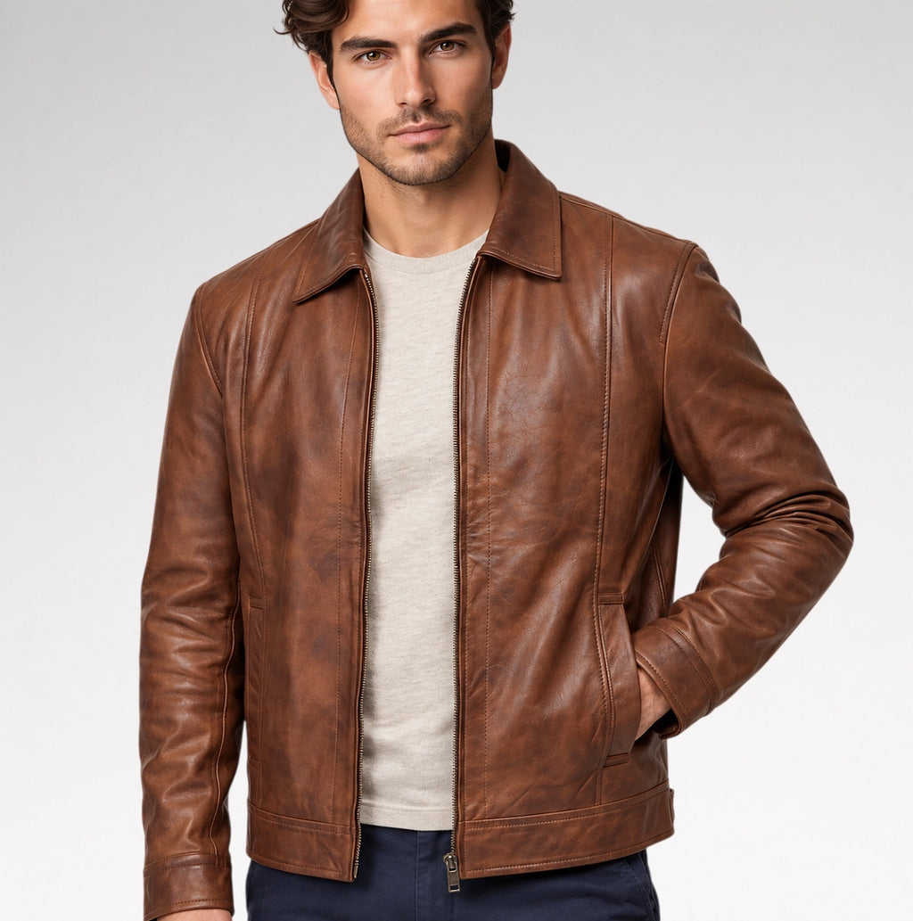 A man wearing a brown leather jacket over a light beige crewneck sweater and dark blue pants, photographed against a clean light gray studio background.