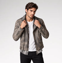 A man wearing a distressed gray leather jacket over a white t-shirt and black pants, posing against a light gray studio background.