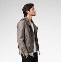 A man wearing a distressed gray suede biker jacket with silver zipper details and patterned lining, styled over a white t-shirt and black pants, photographed against a light gray studio background.