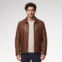 A man wearing a brown leather jacket over a beige T-shirt and dark pants, standing against a light gray studio background.