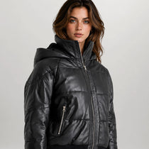A woman wearing a black faux-leather hooded puffer jacket with horizontal quilting and a front zipper, photographed against a light gray studio background.