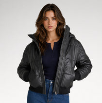A woman modeling a black hooded puffer jacket with a quilted texture, ribbed cuffs and hem, worn over a navy henley top and blue jeans against a light gray studio background.