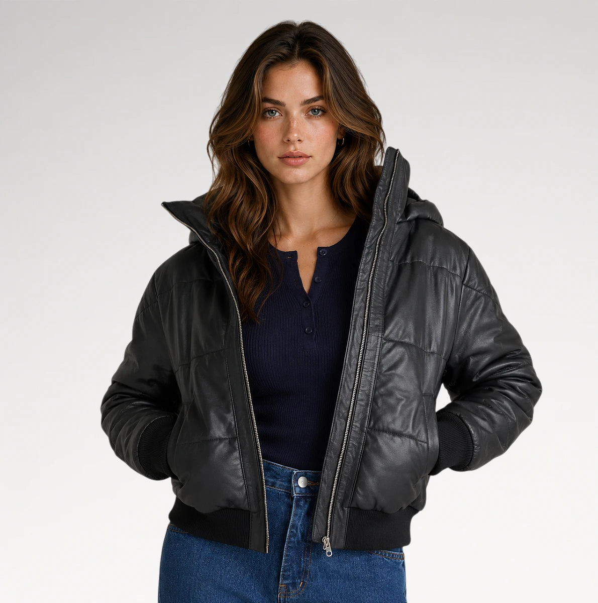 A woman modeling a black hooded puffer jacket with a quilted texture, ribbed cuffs and hem, worn over a navy henley top and blue jeans against a light gray studio background.
