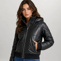 A woman wearing a black faux-leather hooded puffer jacket with a front zipper, ribbed hem and cuffs, paired with blue jeans, photographed against a neutral light gray background.