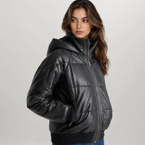 A woman wearing a shiny black hooded puffer jacket with horizontal quilting, photographed against a plain light gray studio background.