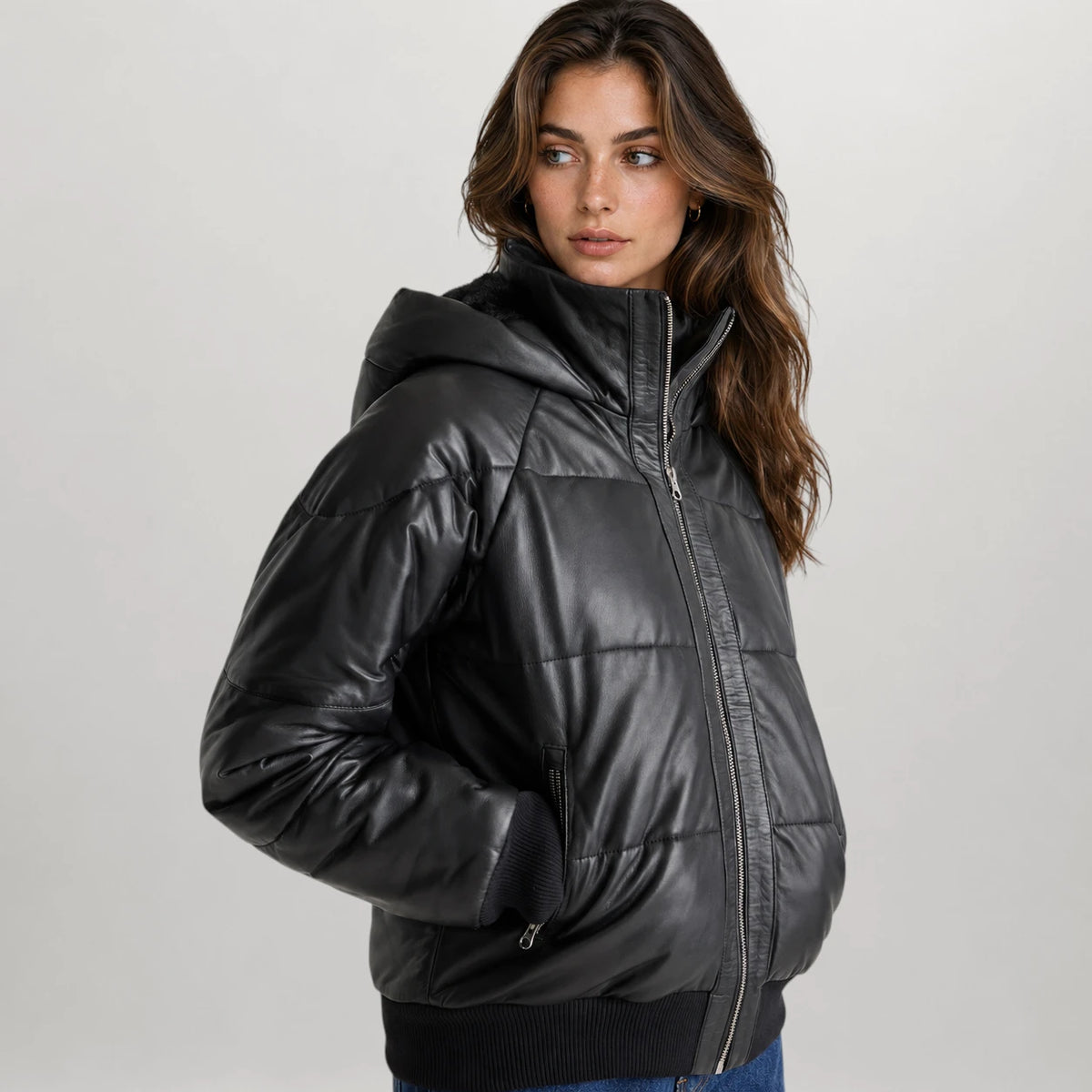 A woman wearing a shiny black hooded puffer jacket with horizontal quilting, photographed against a plain light gray studio background.