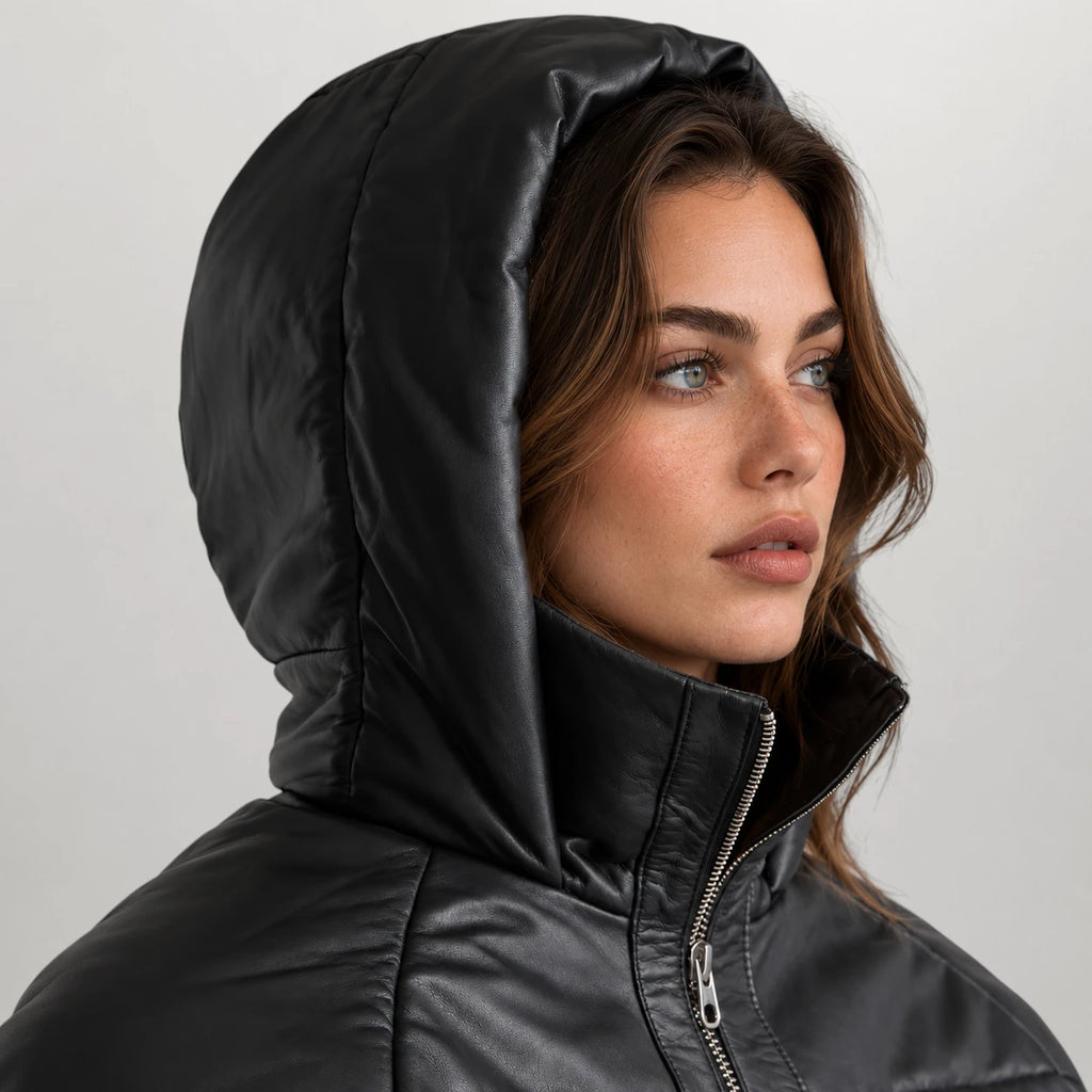 A close-up of a woman wearing a black hooded puffer jacket with a high collar and silver zipper, set against a plain light gray background.