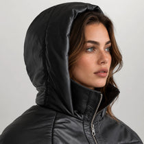 A close-up of a woman wearing a black hooded puffer jacket with a high collar and silver zipper, set against a plain light gray background.