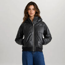 A woman modeling a black quilted puffer jacket with a hood and front zipper, paired with blue jeans, against a light gray studio background.