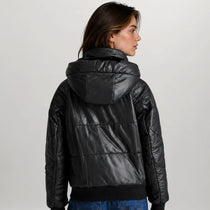 A woman shown from the back wearing a shiny black puffer jacket with a high padded collar and quilted horizontal sections, paired with blue jeans against a light gray studio background.
