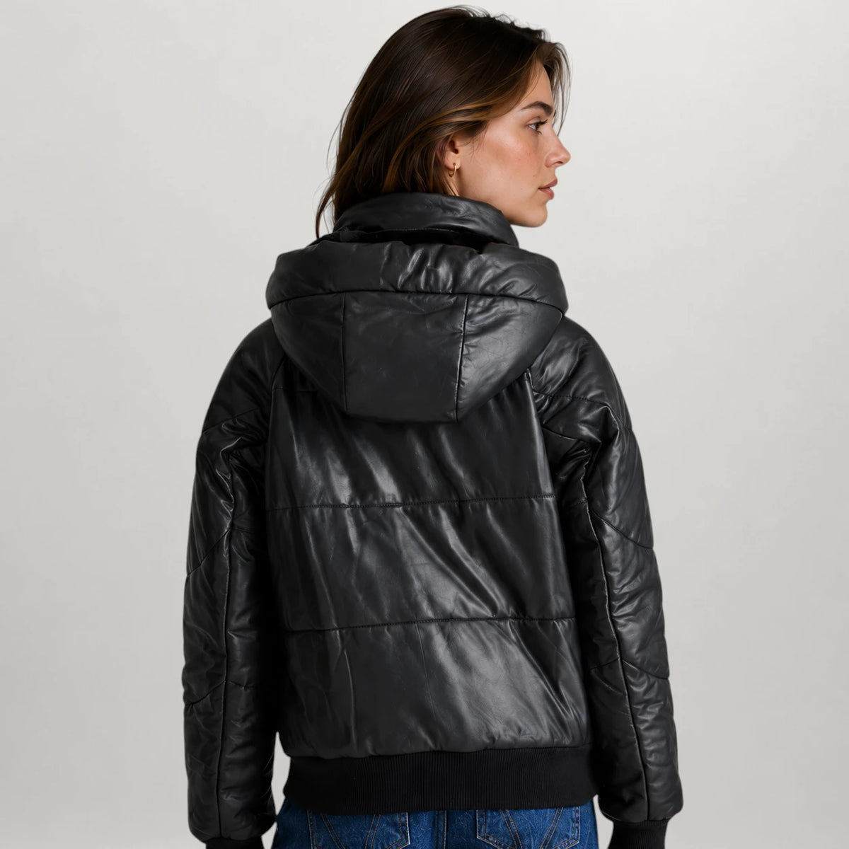 A woman shown from the back wearing a shiny black puffer jacket with a high padded collar and quilted horizontal sections, paired with blue jeans against a light gray studio background.