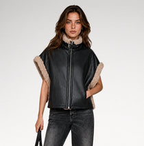A woman modeling a black faux-leather jacket with beige sherpa lining on the collar and sleeve cuffs, paired with dark gray jeans, against a light gray studio background.